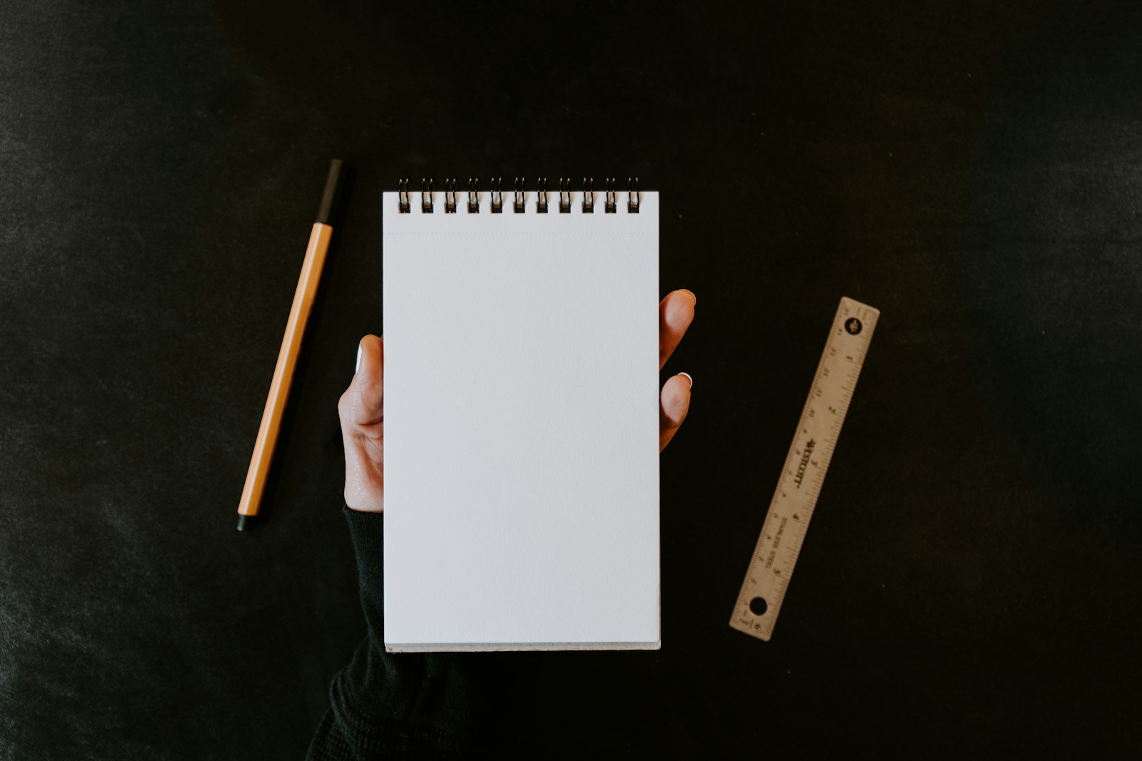 Hand holding a blank sketchpad beside a drawing pen and wooden ruler on a dark surface.